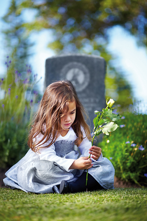 Ein kleines Mädchen sitzt vor dem Grab mit einer weißen Rose in der Hand.